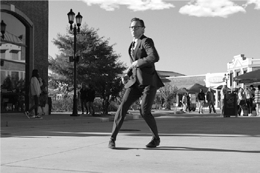 Chuck, in a suit, is mid-step with one leg extended and slightly blurred, indicating movement in his tap dance on a paved plaza. His arms are swinging.