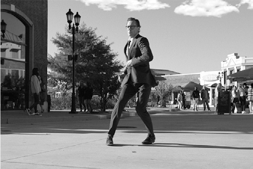 Chuck is mid-step with his right leg extended forward and left leg bent back, arms bent, on a paved plaza. His arms are swinging.