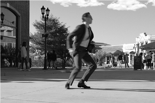 Chuck’s legs bent sideways, a rapid movement, arms extended sideways, body leans forward, on a outdoor plaza.