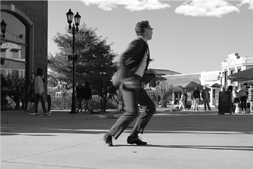 Chuck’s legs bent in a fast small steps, arms held out, body moving rapidly sideways, on a outdoor plaza.